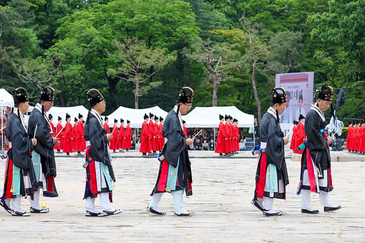 Homens de robe preto e vermelho e chapéu preto caminham em fila pelo pátio de pedra, com outro grupo de robe vermelho atrás.