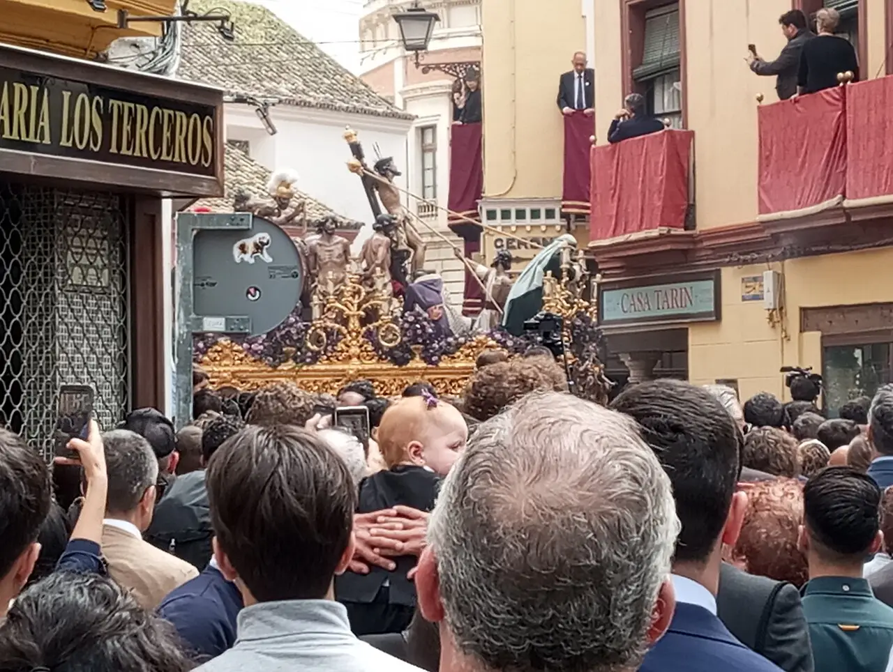 Vista por trás da multidão, um passo dourado com figuras segue por uma rua estreita, e à esquerda o letreiro diz Los Terceros.