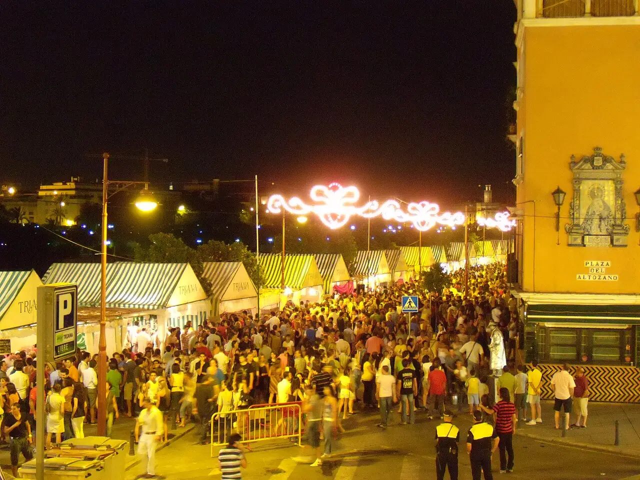 À noite, a multidão enche a rua entre barracas de lona listrada, sob luzes em forma de coração, com um prédio amarelo à direita.