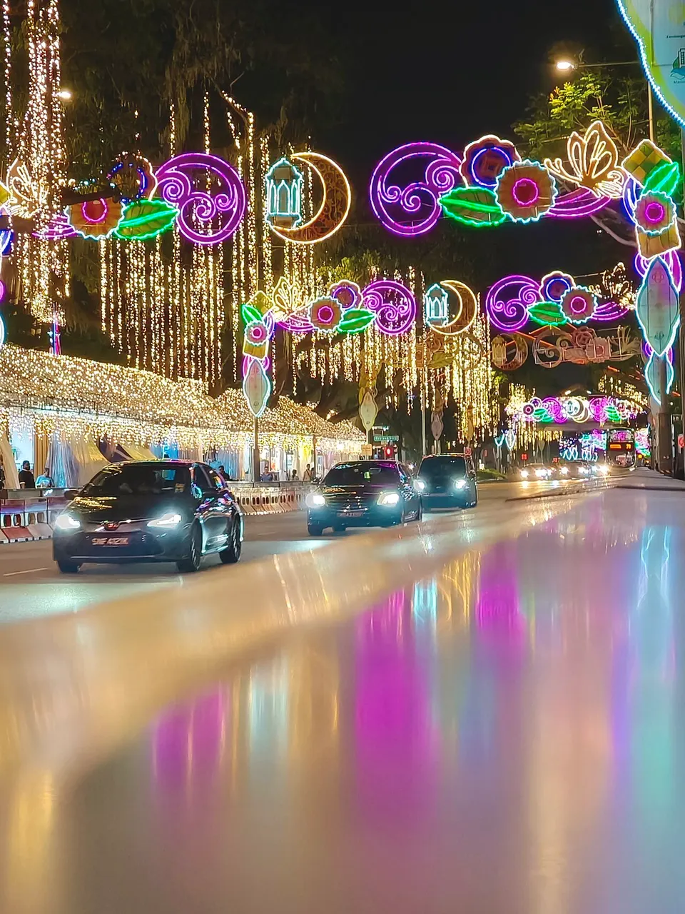 Rua de Geylang Serai com crescentes, lanternas e arabescos luminosos sobre carros e reflexos coloridos no pavimento molhado.