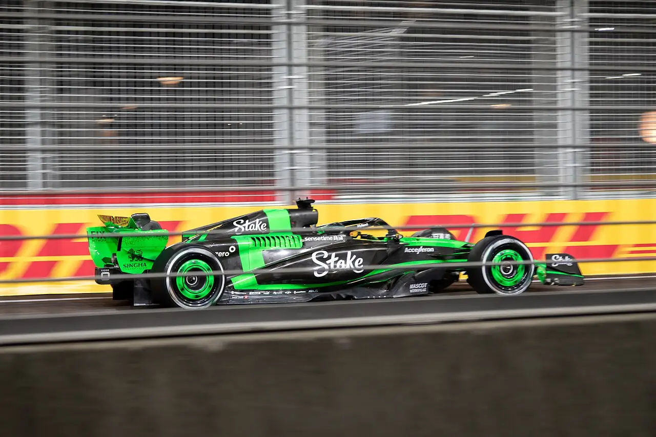 Um carro de fórmula verde e preto passa junto à grade, com a barreira amarela da DHL atrás.