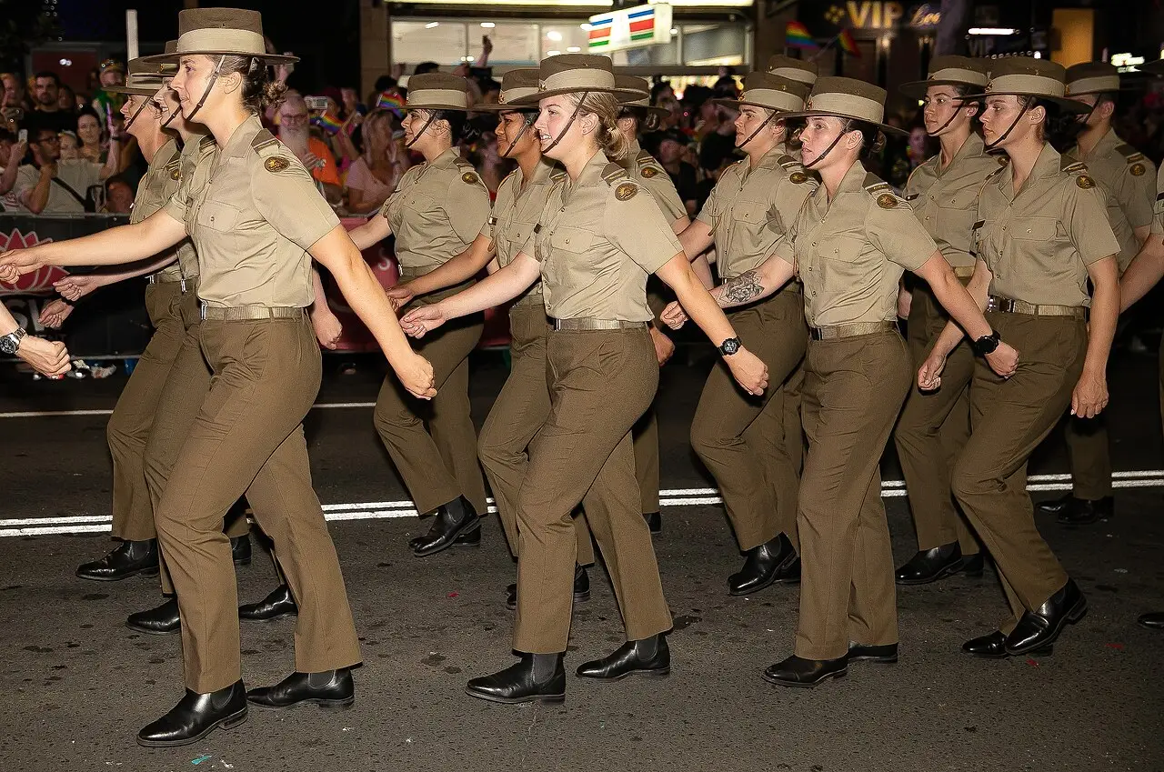 Soldados de uniforme cáqui e chapéu marcham em fila na rua, à noite, com o público atrás.