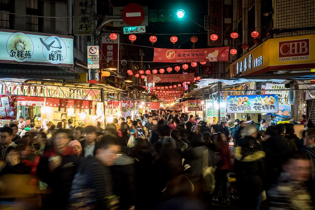 À noite, a rua do mercado está cheia de gente, com lanternas vermelhas penduradas e barracas iluminadas dos dois lados.