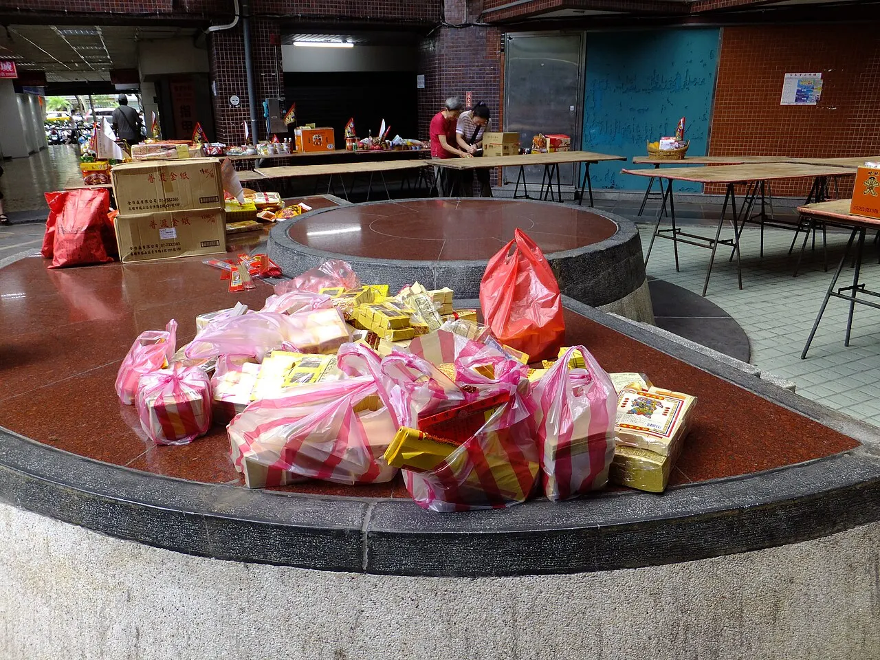 Pacotes de papel ritual e sacolas vermelhas e rosas empilhados sobre uma plataforma de pedra, com mesas de oferendas ao fundo.