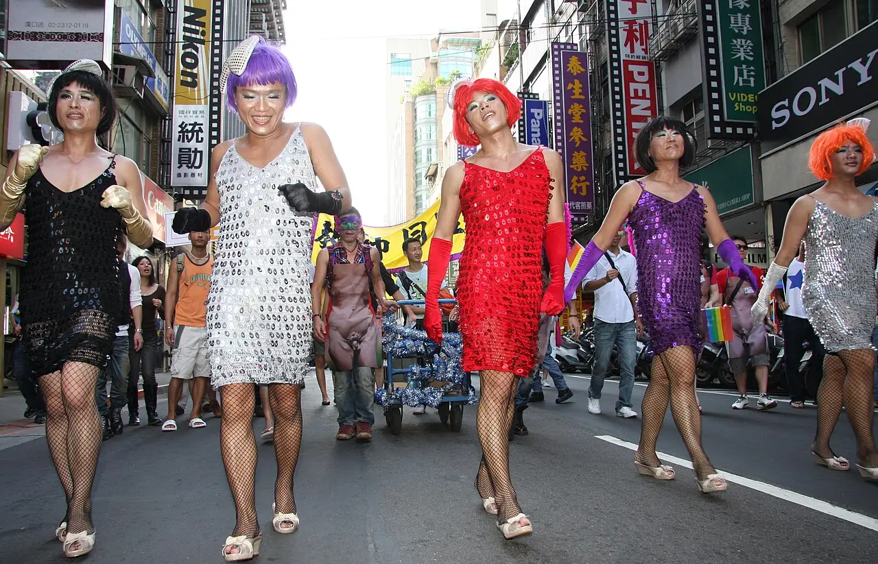 Cinco pessoas de vestido curto e peruca colorida marcham pela rua, com a multidão atrás e lojas dos dois lados.