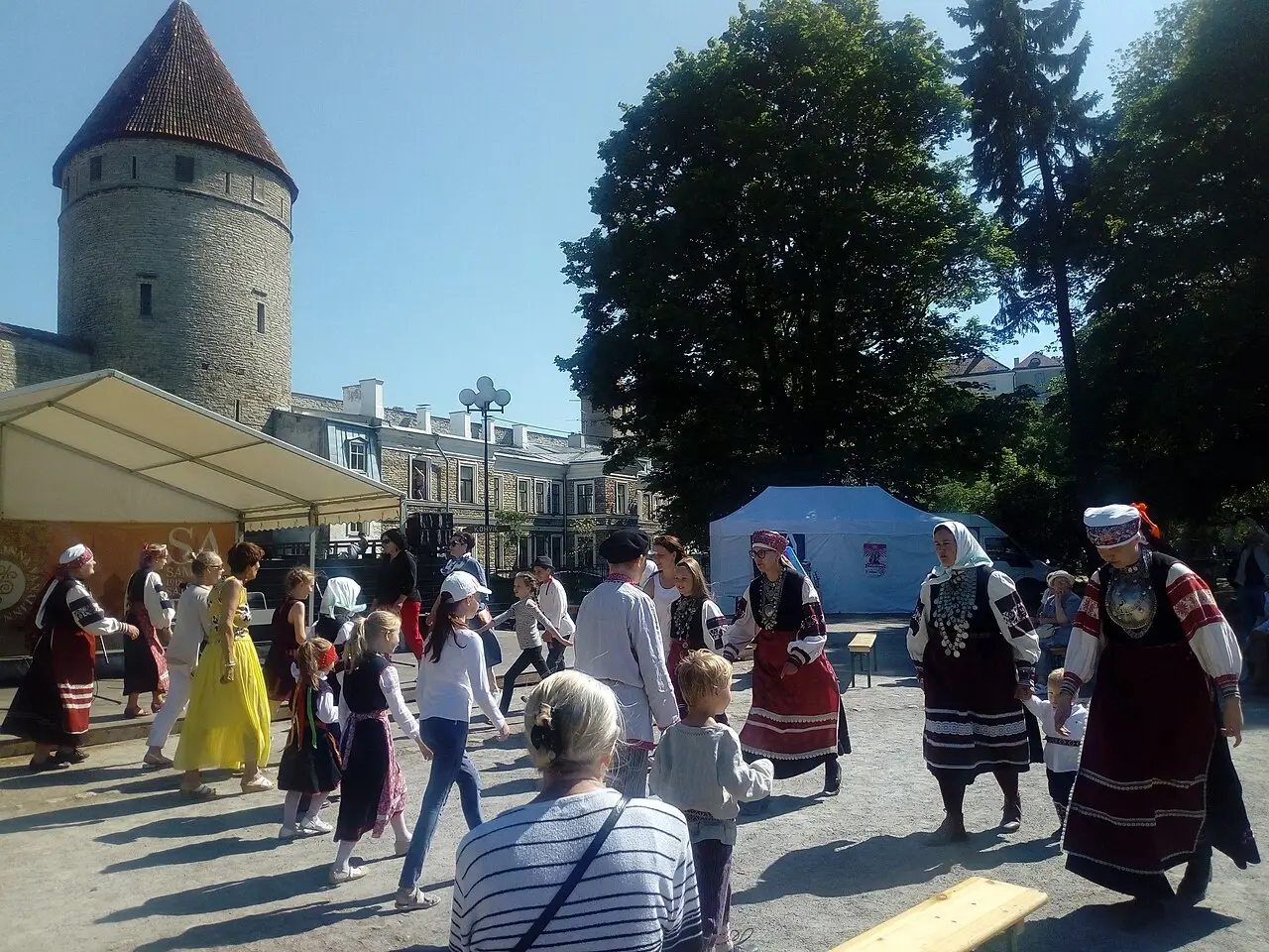 Pessoas de traje tradicional, com saia vermelha e colete, dançam na praça, com uma torre redonda de pedra à esquerda e o público em volta.
