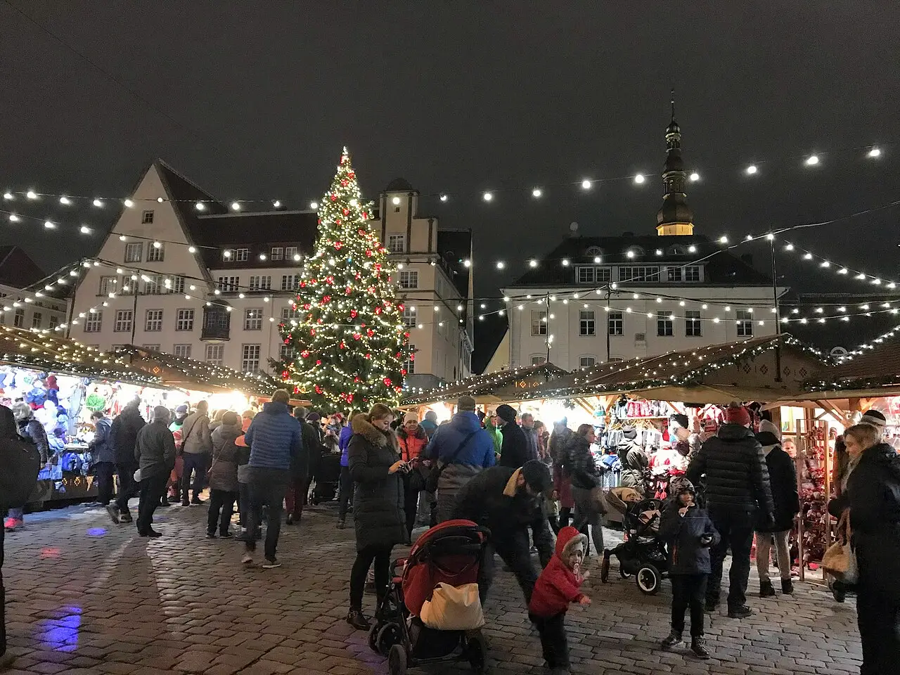 À noite, pessoas passam entre as barracas e uma árvore de Natal iluminada, sob fios de lâmpadas, com as casas da praça atrás.