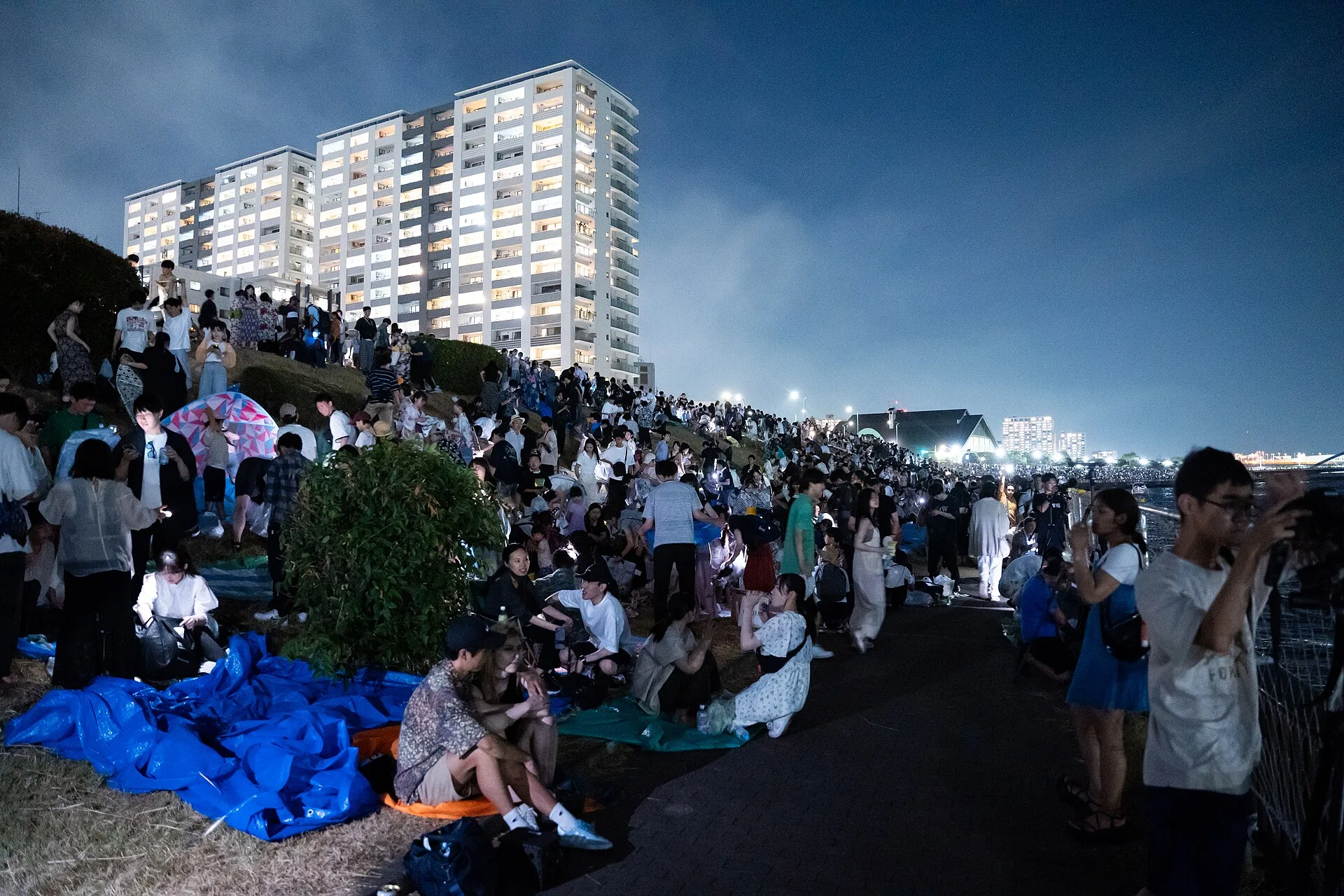 Gente sentada e em pé ao longo da margem, com torres de apartamentos atrás, na festa de fogos de 2023.
