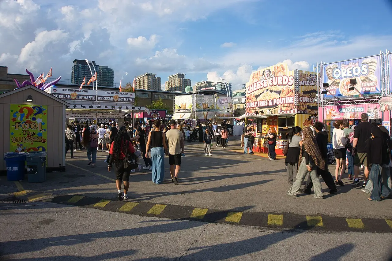 Personas caminan entre los puestos de la feria, con un quiosco de madera a la izquierda y torres de la ciudad al fondo.