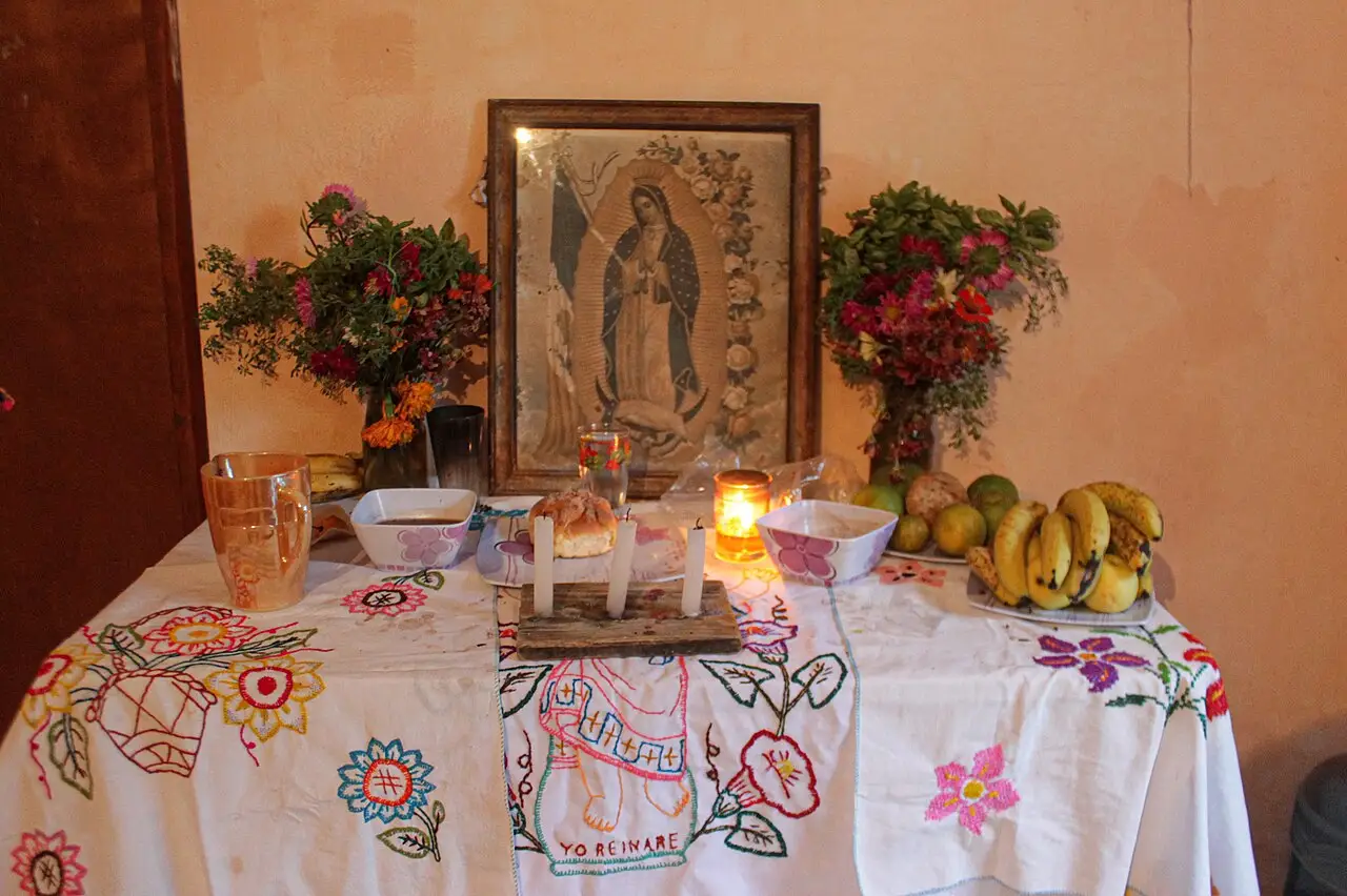 A mesa, com toalha branca bordada, tem velas acesas, bananas, flores e um quadro da Virgem ao fundo.