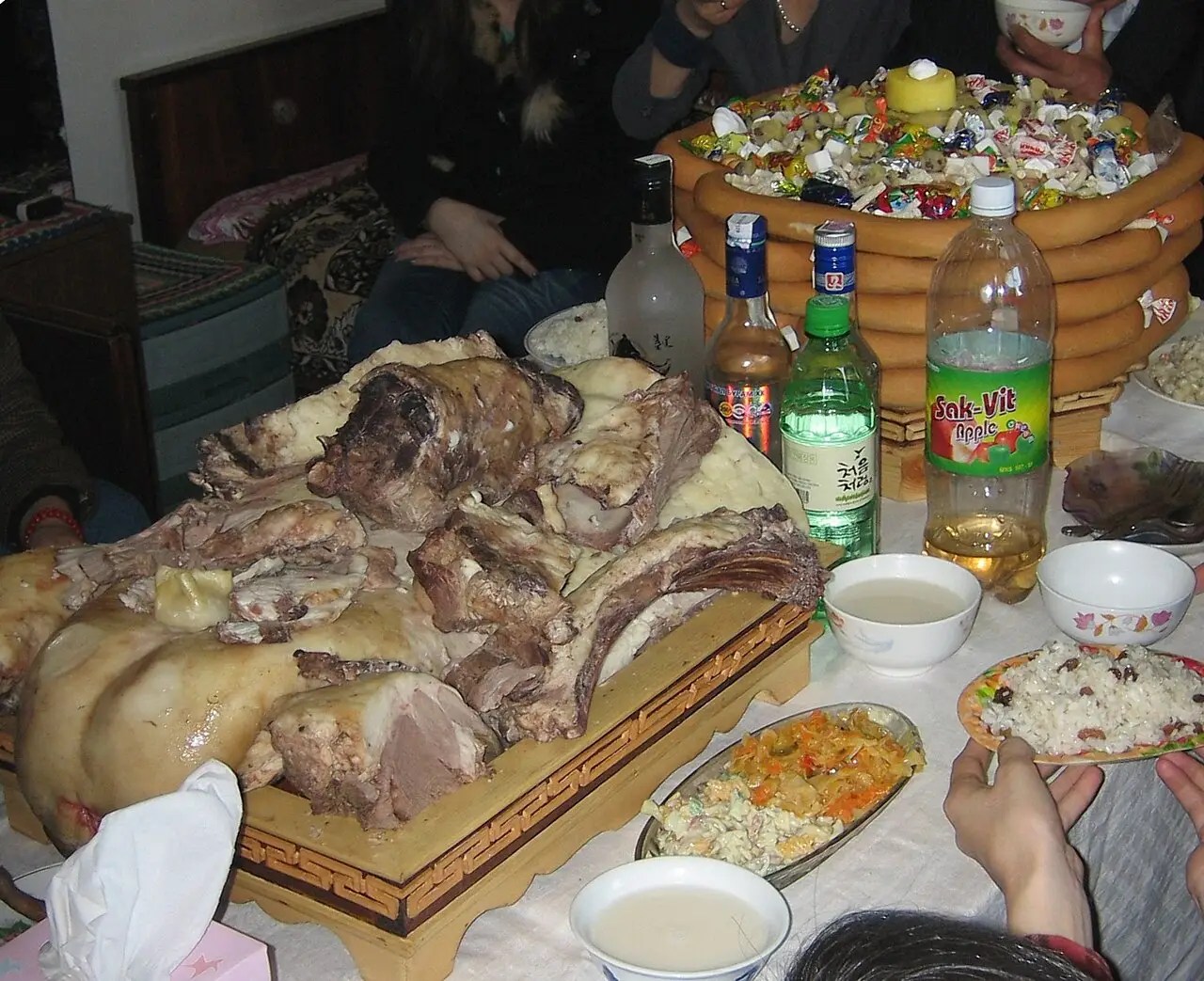A mesa reúne um grande corte de carne, garrafas, tigelas e uma torre de biscoitos coloridos, com pessoas sentadas em volta.