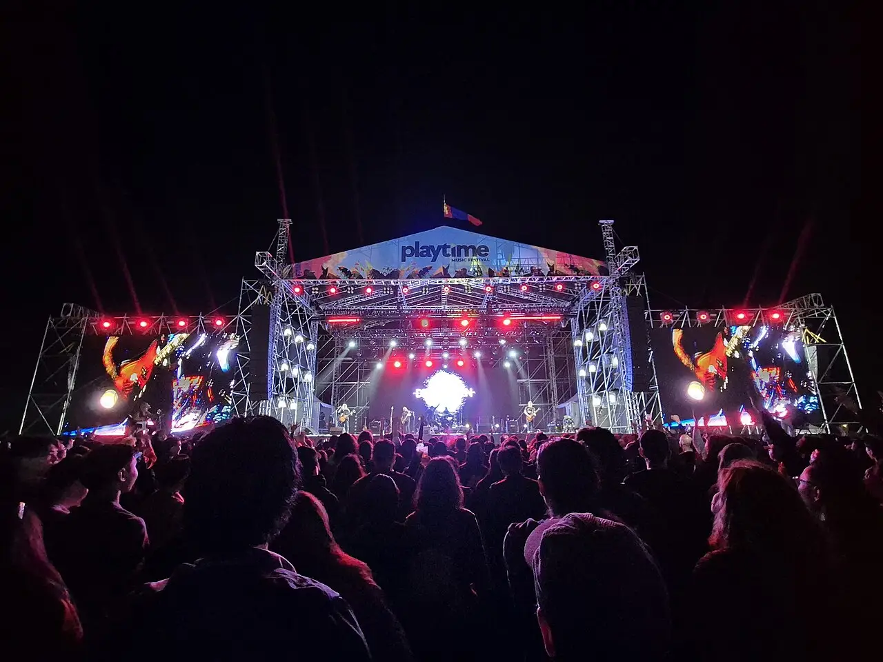 De costas, o público olha o palco iluminado, com telões dos dois lados e o letreiro playtime no alto.