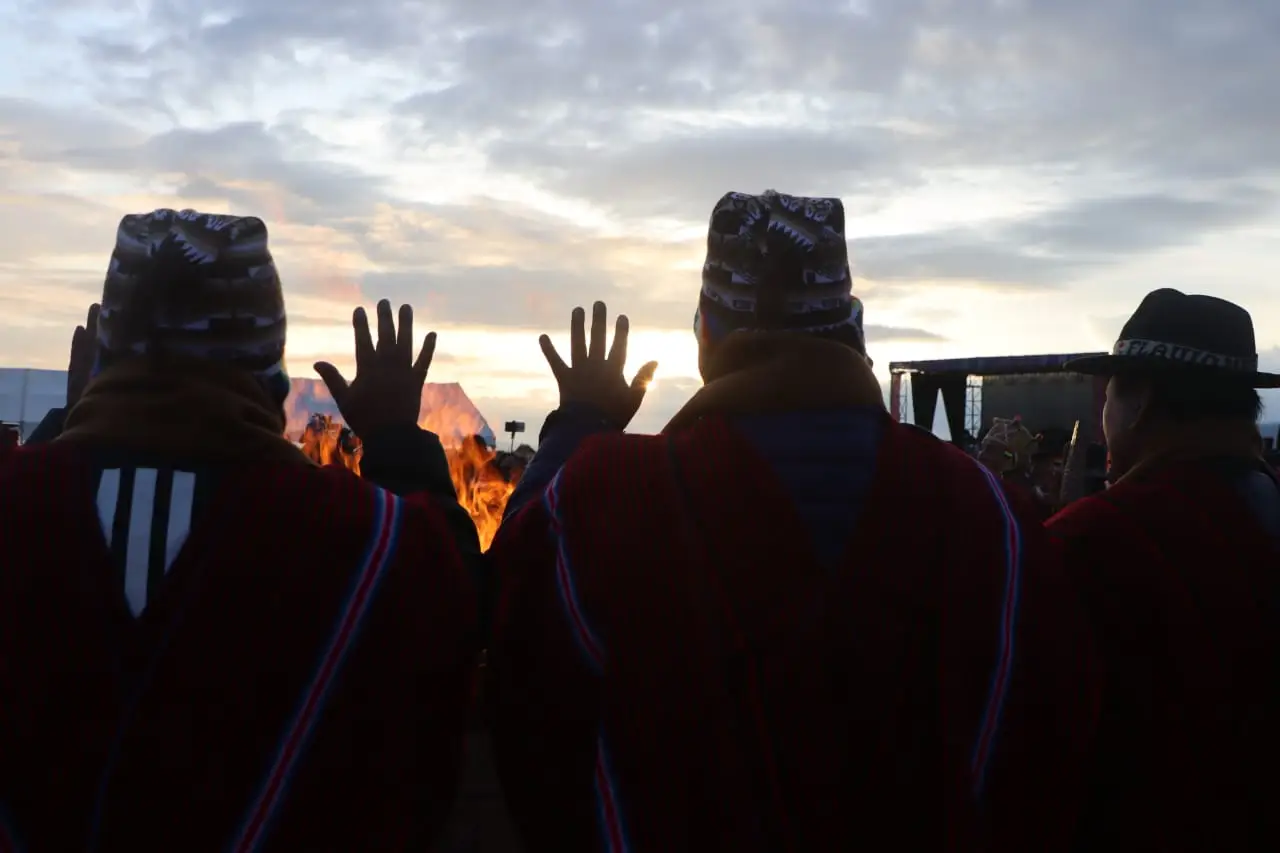 De costas, pessoas de poncho vermelho e gorro estampado erguem as mãos abertas para a luz no horizonte, com uma fogueira baixa à frente.