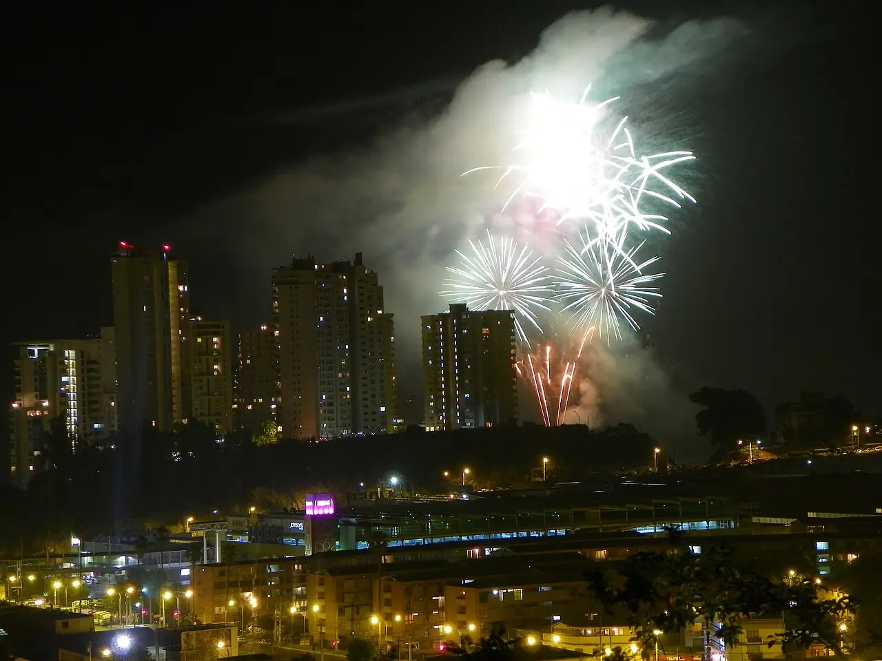 Fogos brancos e vermelhos estouram acima dos prédios altos, e a cidade fica acesa embaixo.