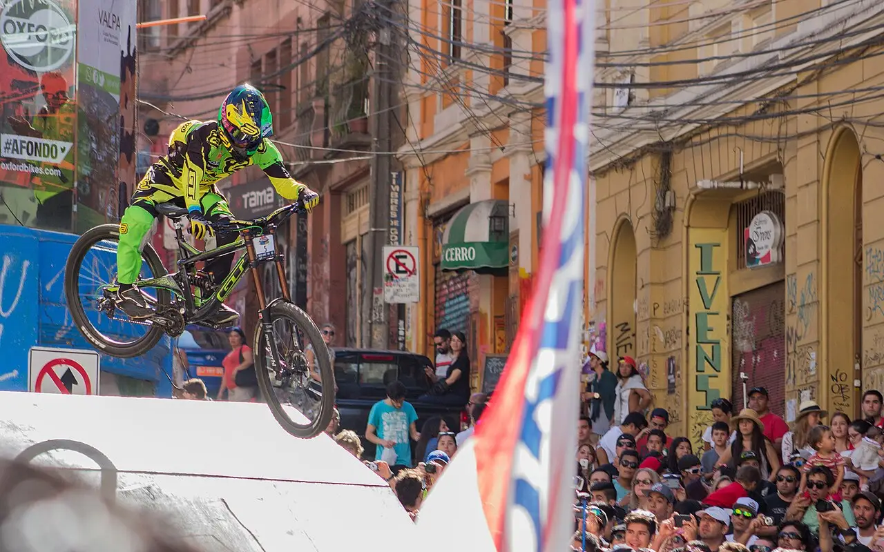 Um ciclista de verde e amarelo está no ar com a bicicleta, sobre uma rampa, e o público acompanha da calçada.