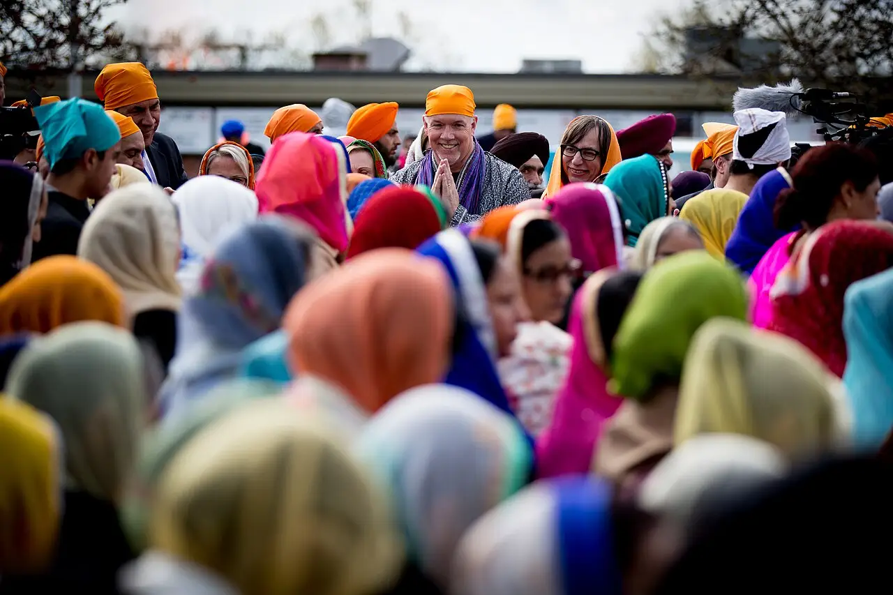 Uma multidão de turbantes e lenços coloridos se aperta, e um homem de turbante laranja sorri no meio.