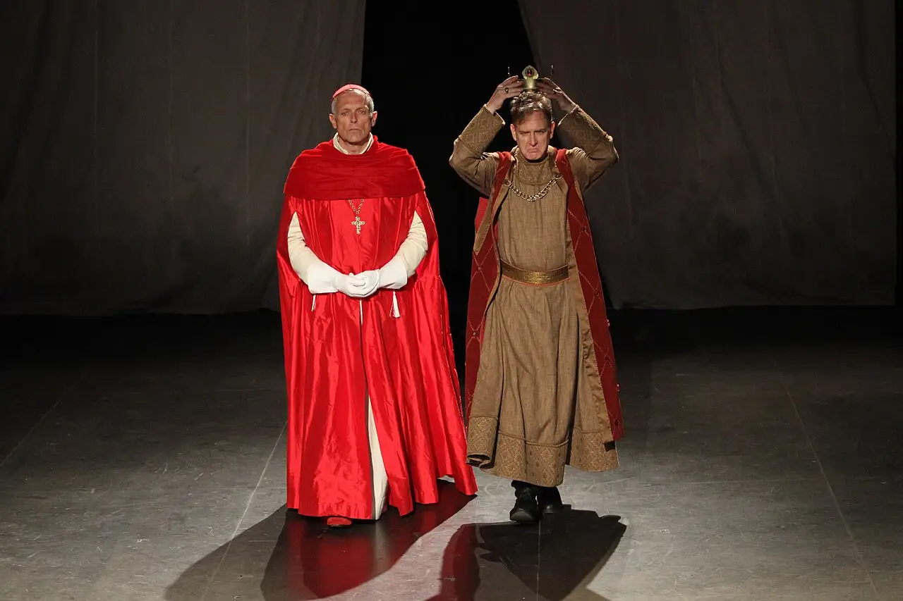 No palco, um homem de manto vermelho está ao lado de outro, de túnica marrom e capa, que leva as duas mãos à cabeça.