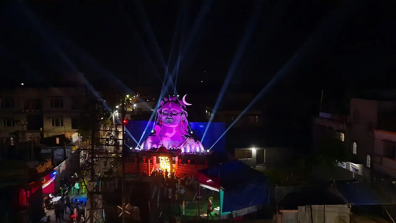 À noite, um rosto enorme de Shiva, iluminado de roxo, fica no alto de um palco, com feixes de luz e prédios em volta.