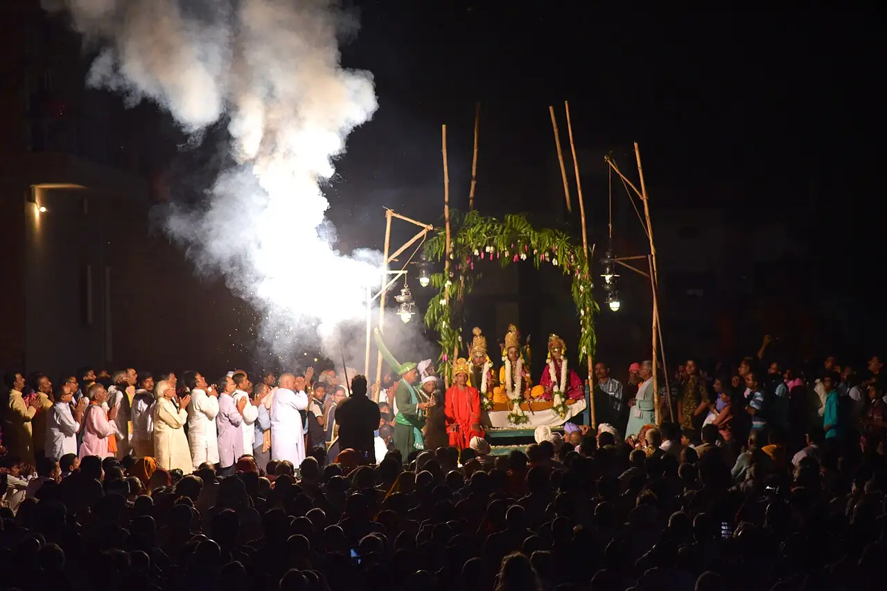 À noite, o público vê um palco com um arco de ramos, fumaça branca e pessoas de rosa, branco e vermelho.