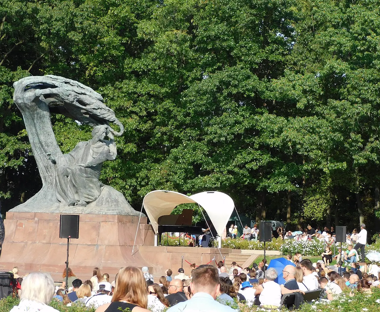 O público sentado olha um palco com toldo branco, ao lado do monumento de Chopin, com as árvores atrás.