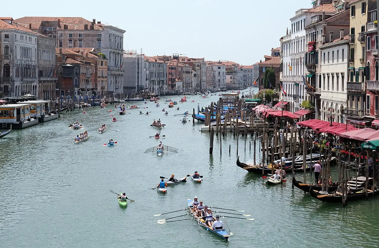 Vários barcos a remo descem o canal, entre os prédios das duas margens, com gôndolas atracadas à direita.