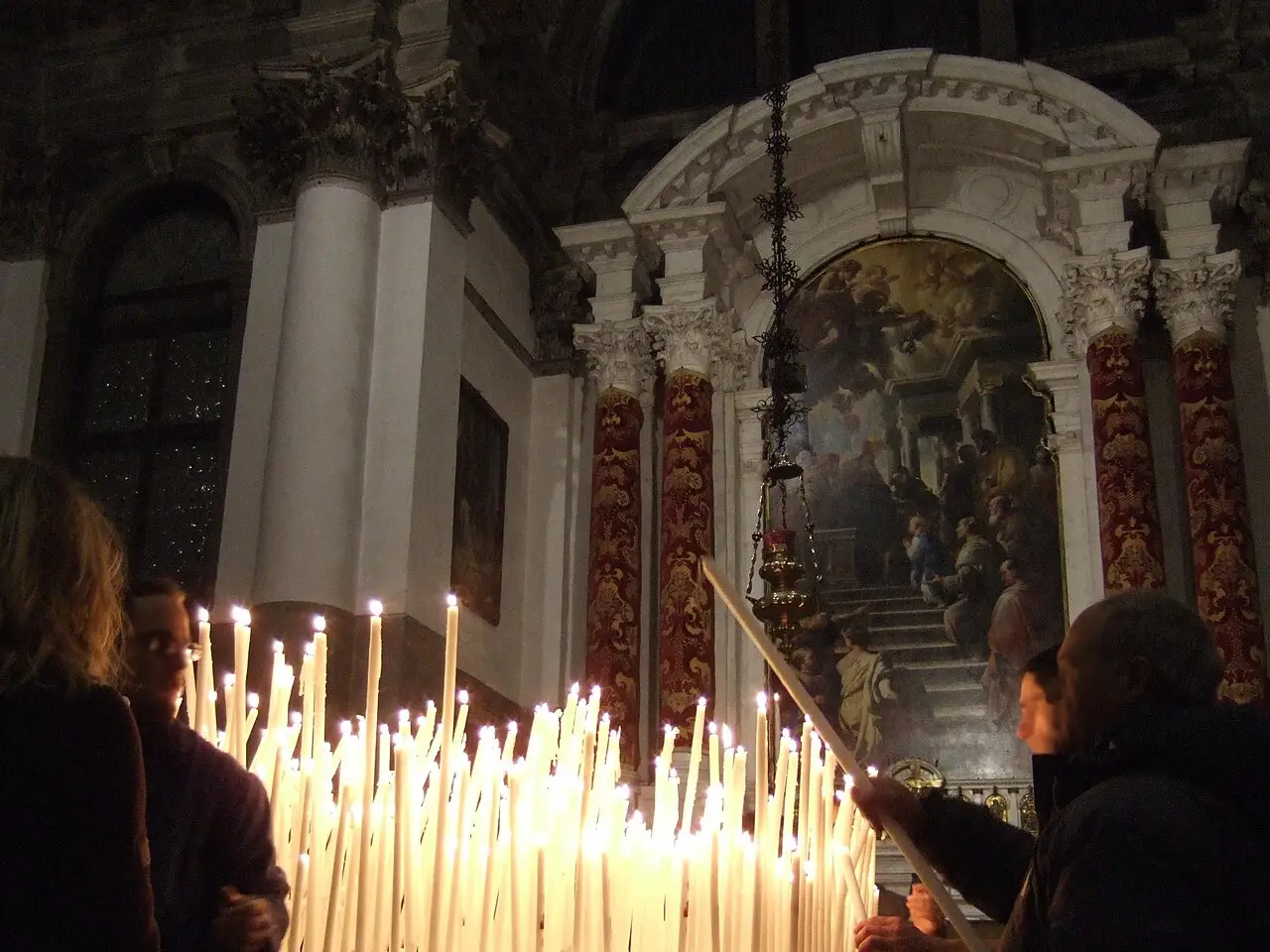Dentro da igreja, uma pessoa acende uma vela num suporte cheio de velas, com um altar pintado ao fundo.