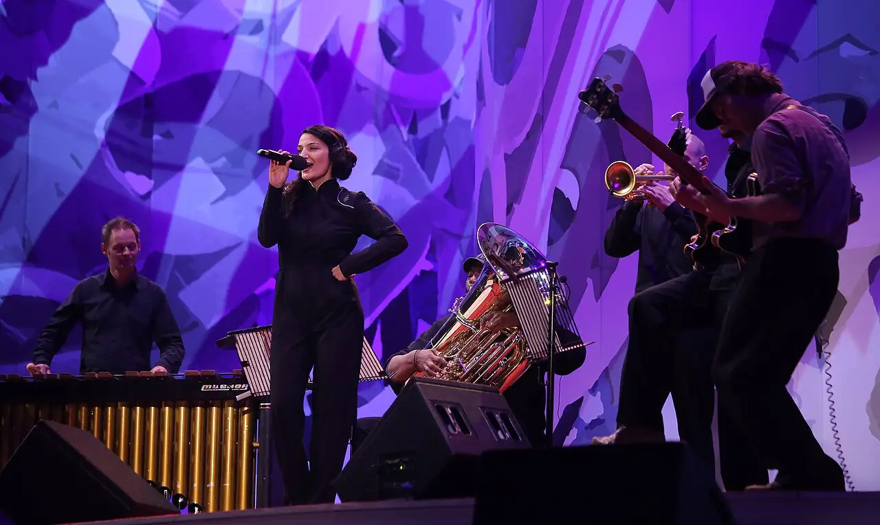 No palco, uma cantora de macacão preto segura o microfone, com trompete, guitarra e marimba, diante de um fundo roxo.