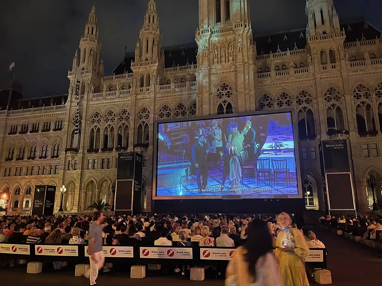 At night, the seated audience watches a show on the large screen, in front of the illuminated city hall.