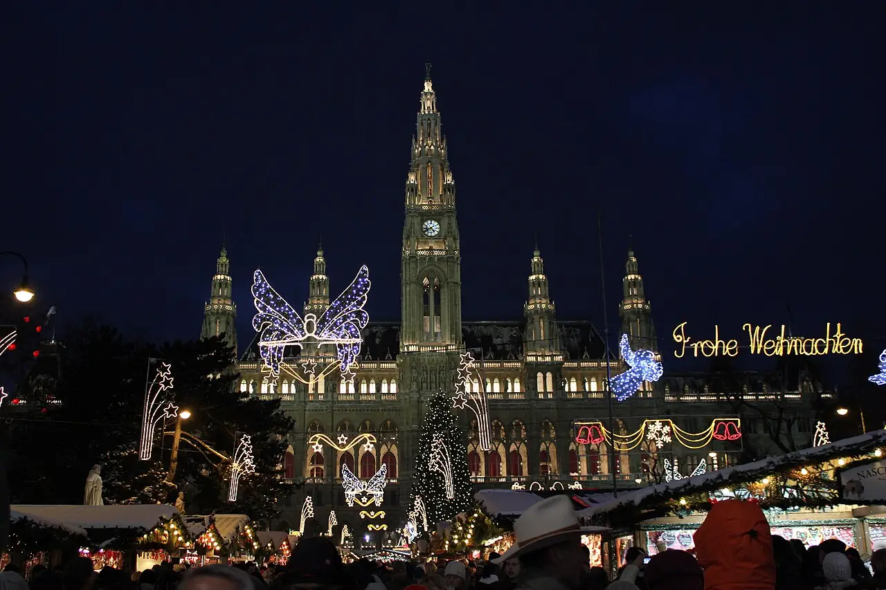 At night, stalls and people stand in front of the city hall, with a tree, light butterflies and the Frohe Weihnachten sign.