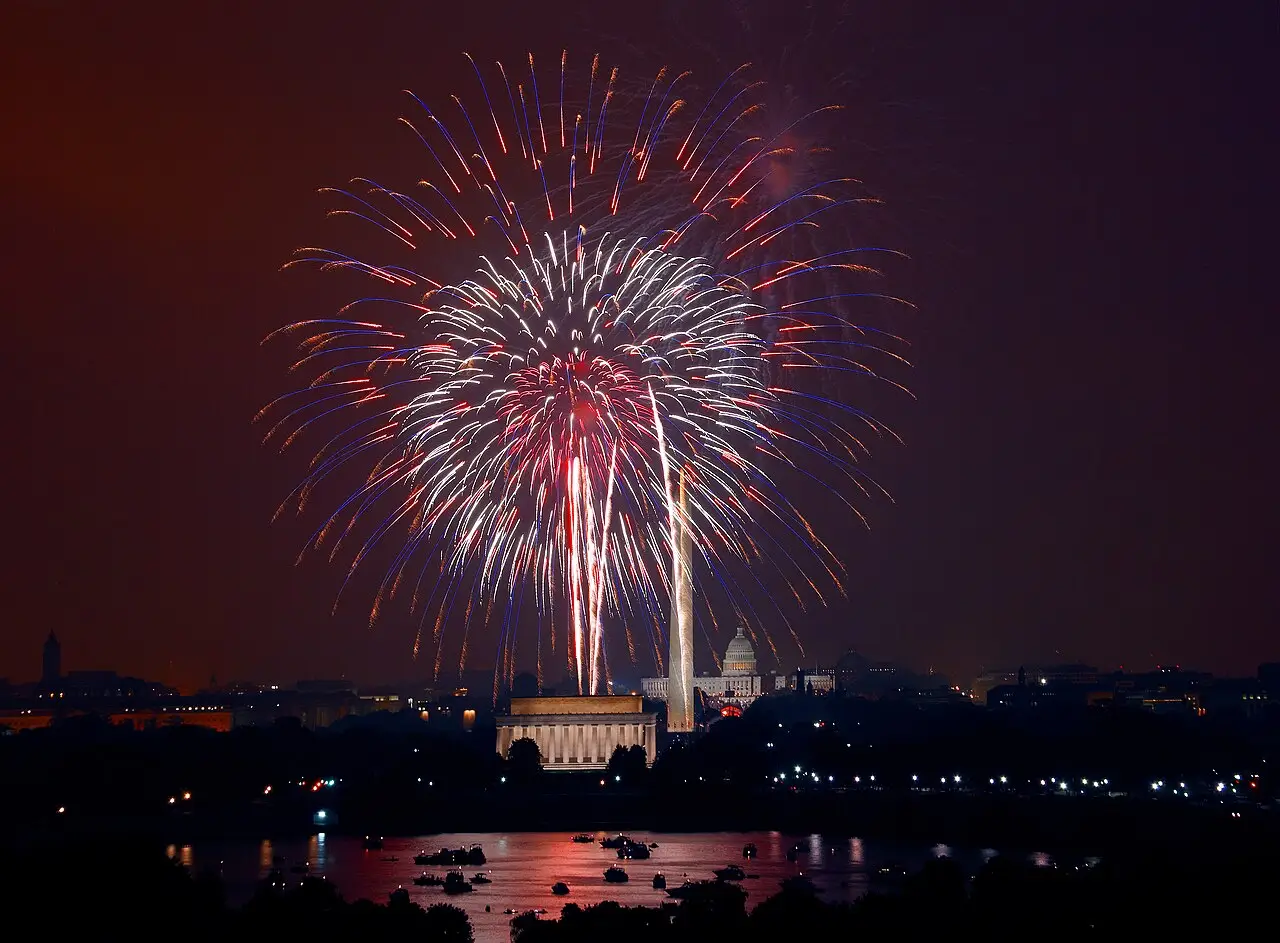 Fogos vermelhos e brancos estouram sobre o Monumento a Washington, e a água reflete as luzes.