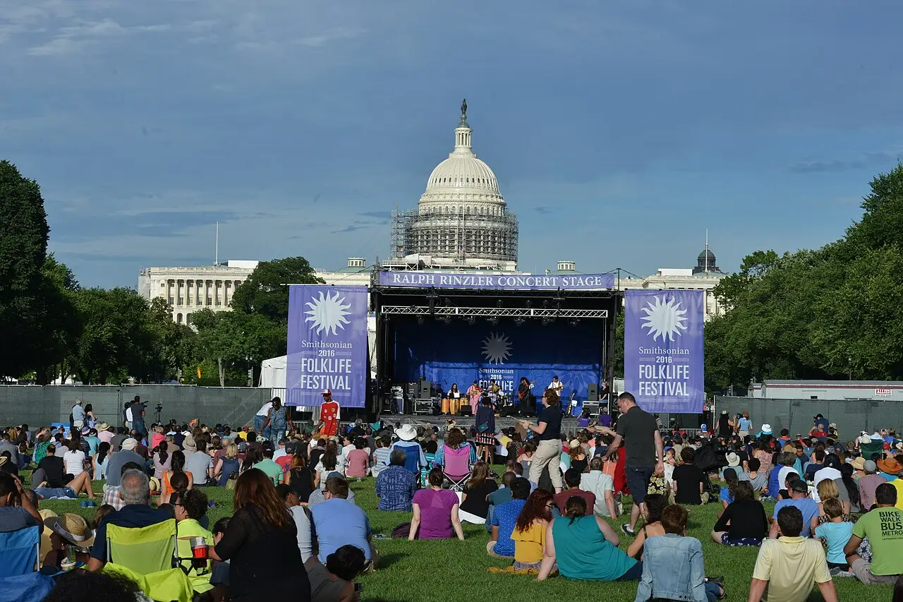 O público sentado na grama olha um palco com faixas do Folklife Festival, e a cúpula do Capitólio fica atrás.