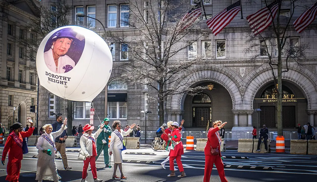 No desfile, pessoas de terno vermelho e chapéu caminham ao lado de um balão branco com um retrato, e bandeiras americanas pendem dos prédios.