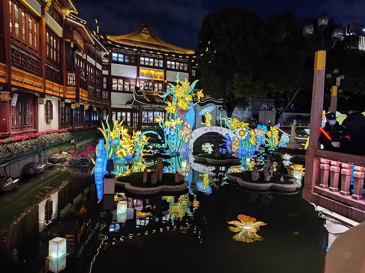 À noite, lanternas em forma de flores e de lótus iluminam o lago, entre prédios de telhado curvo e uma ponte.
