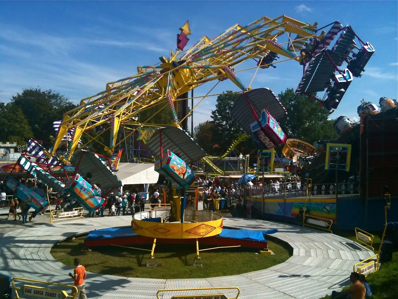 Um brinquedo de feira, com gôndolas no alto de um braço, gira num pátio, com gente em volta.