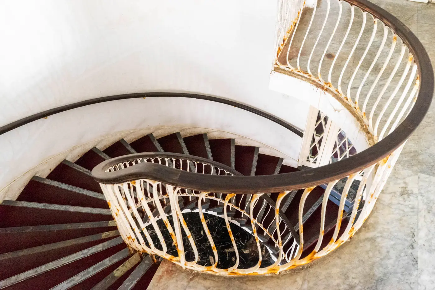 Escada em caracol com corrimão branco e ferrugem, degraus escuros e parede branca.