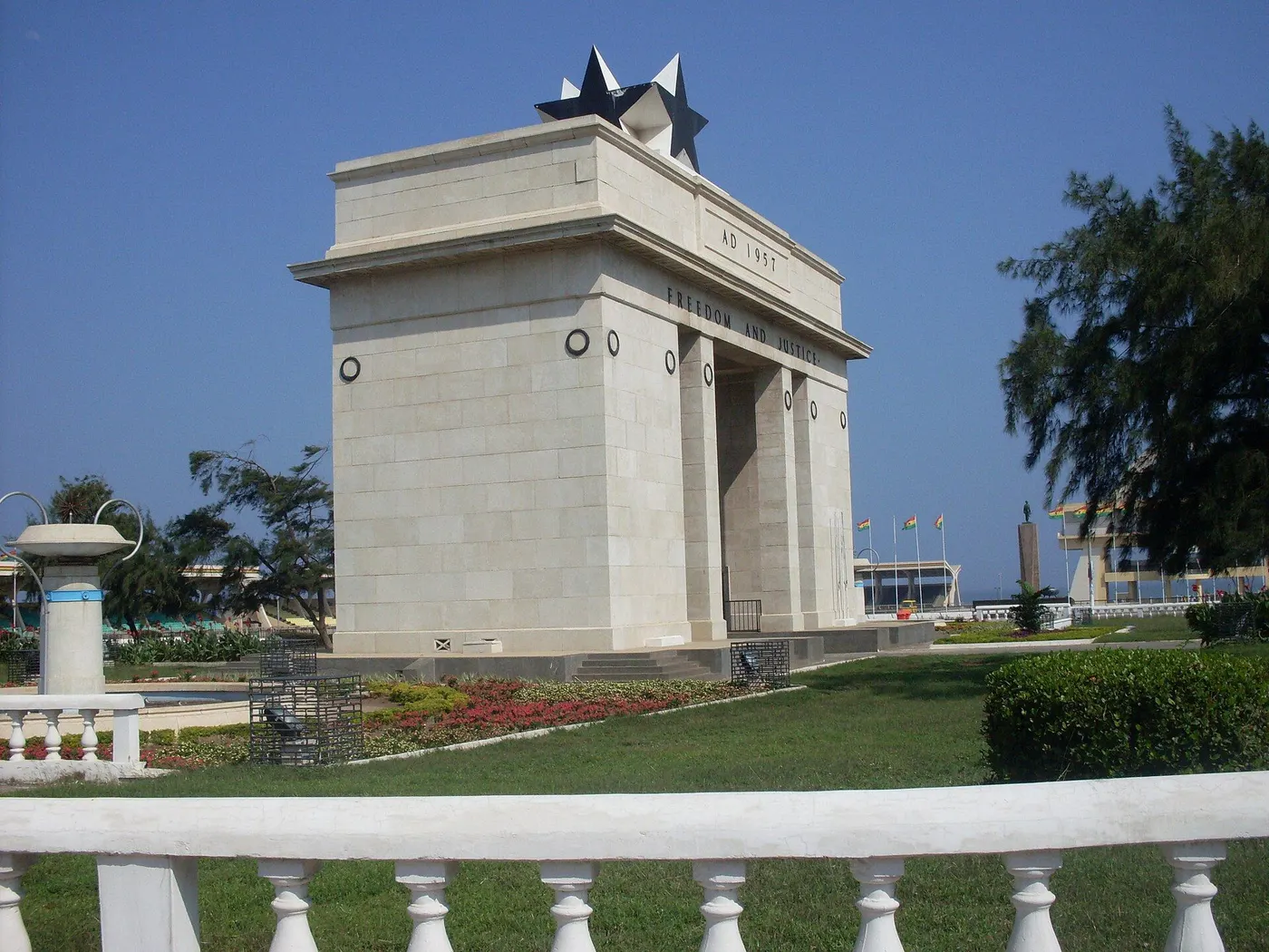 Arco de pedra clara com uma estrela negra no topo, jardim na frente e o mar ao fundo.