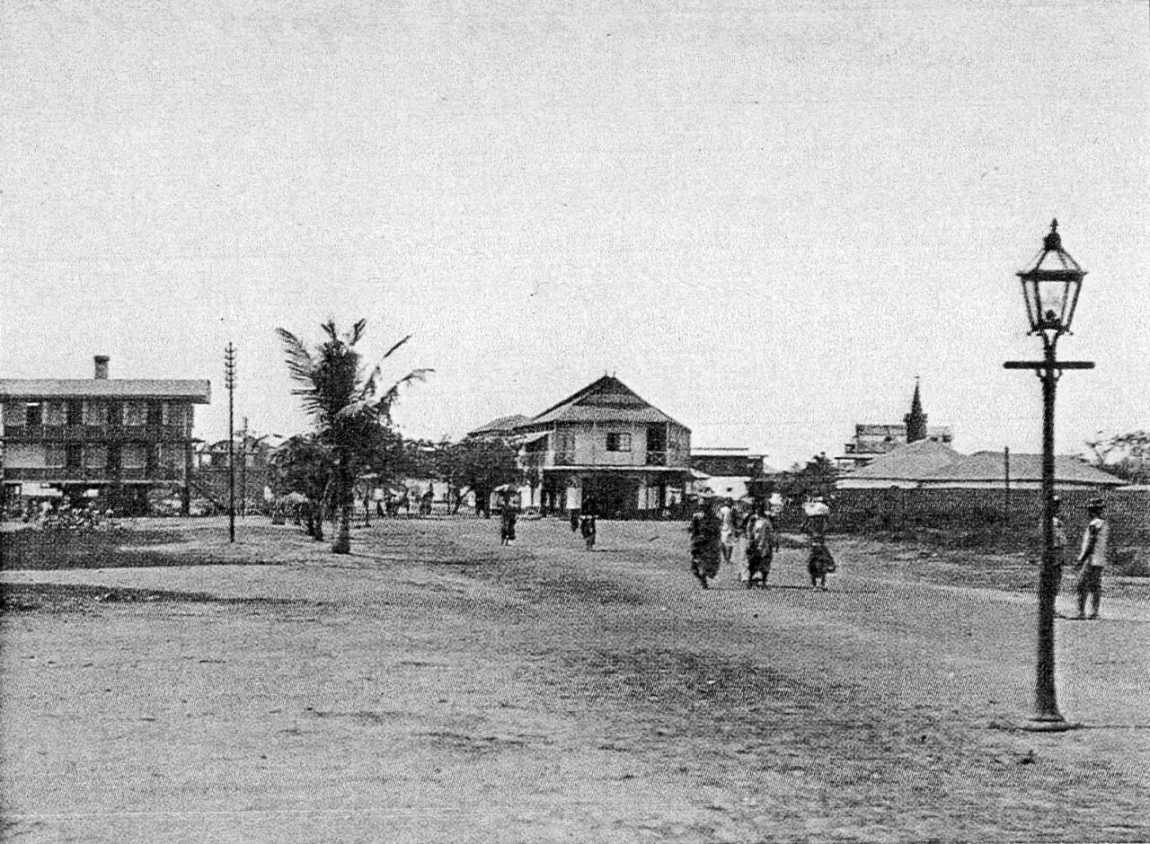 Fotografia em preto e branco de uma rua larga de terra, com sobrados de varanda, um lampião em primeiro plano, pessoas a pé e uma palmeira.