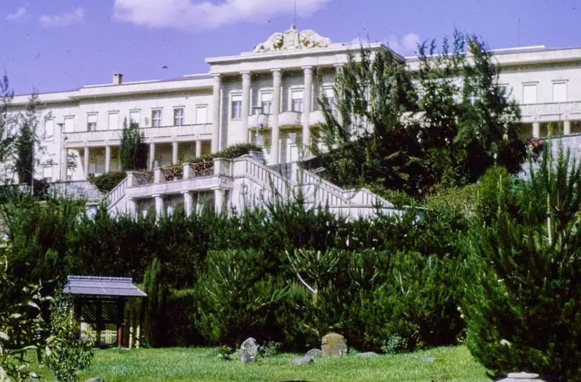Palácio claro com colunas ao centro, escada dupla e jardim de árvores escuras.