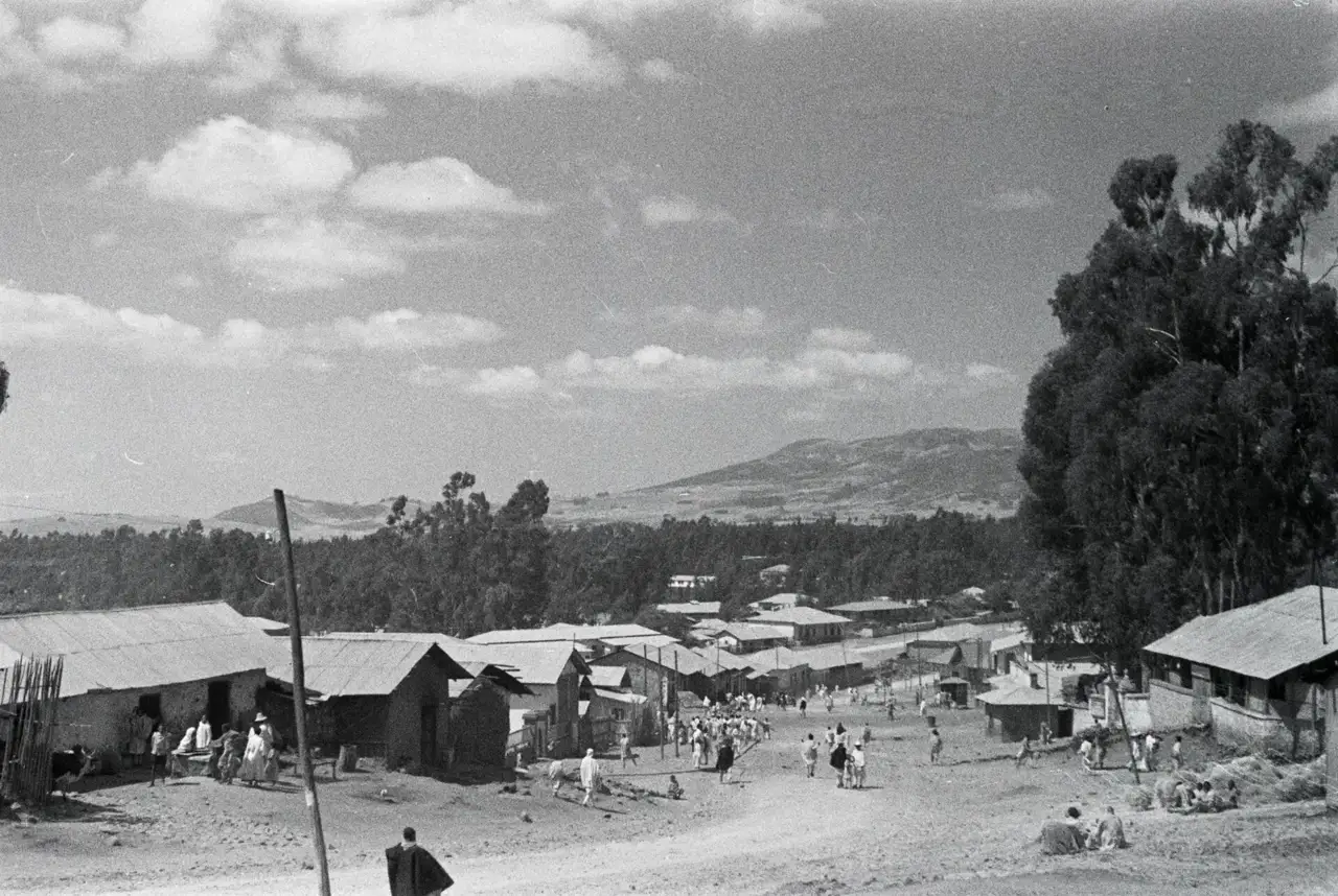 Fotografia em preto e branco de uma rua de terra em Adis Abeba, com pessoas em trajes brancos tradicionais, casas de telhado de zinco, eucaliptos e colinas ao fundo.