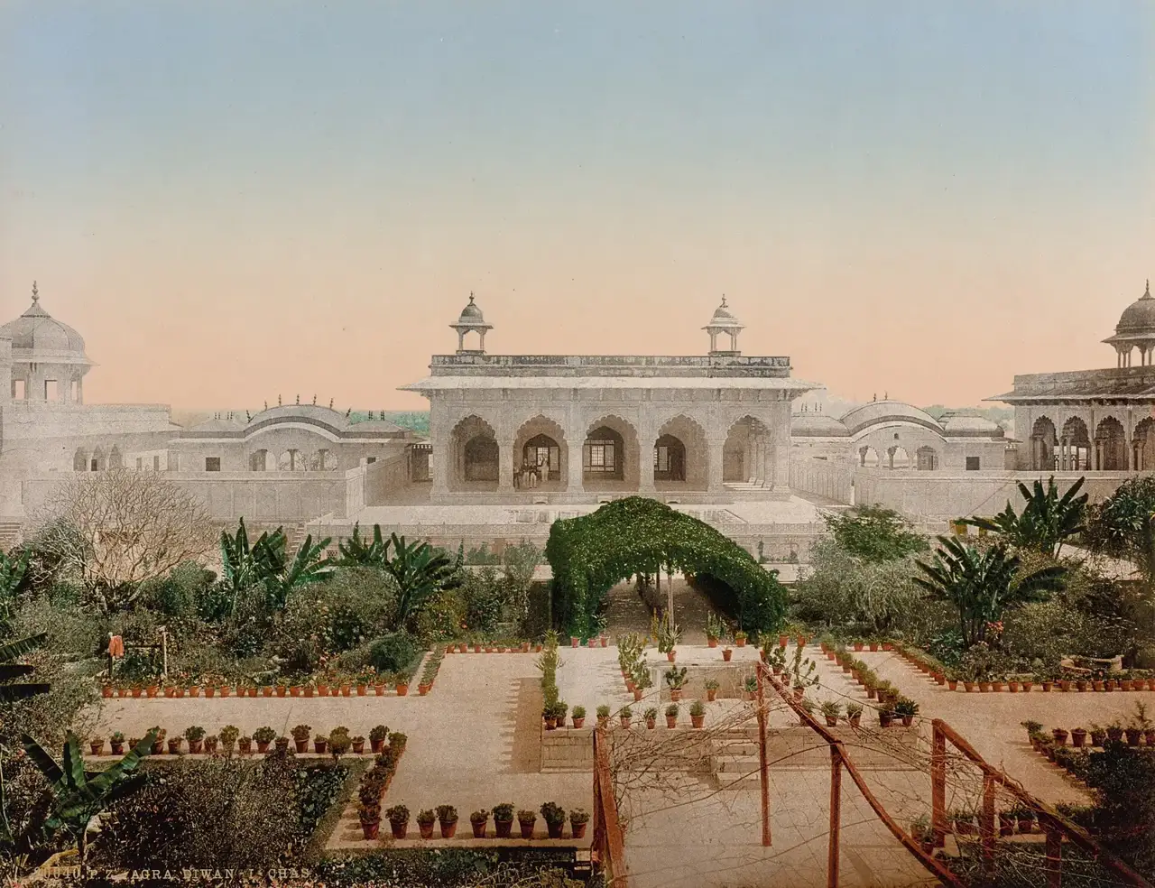 Fotocromo colorido do Diwan-i-Khas, pavilhão de mármore com arcos dentro do Forte de Agra, visto por trás de um jardim com canteiros e vasos de plantas em fileiras.