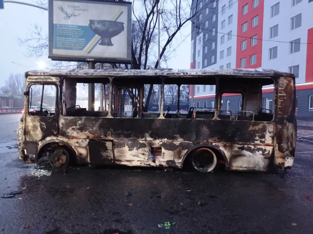 Ônibus sem vidros e com a lataria queimada, parado numa rua molhada, com um prédio e um painel atrás.