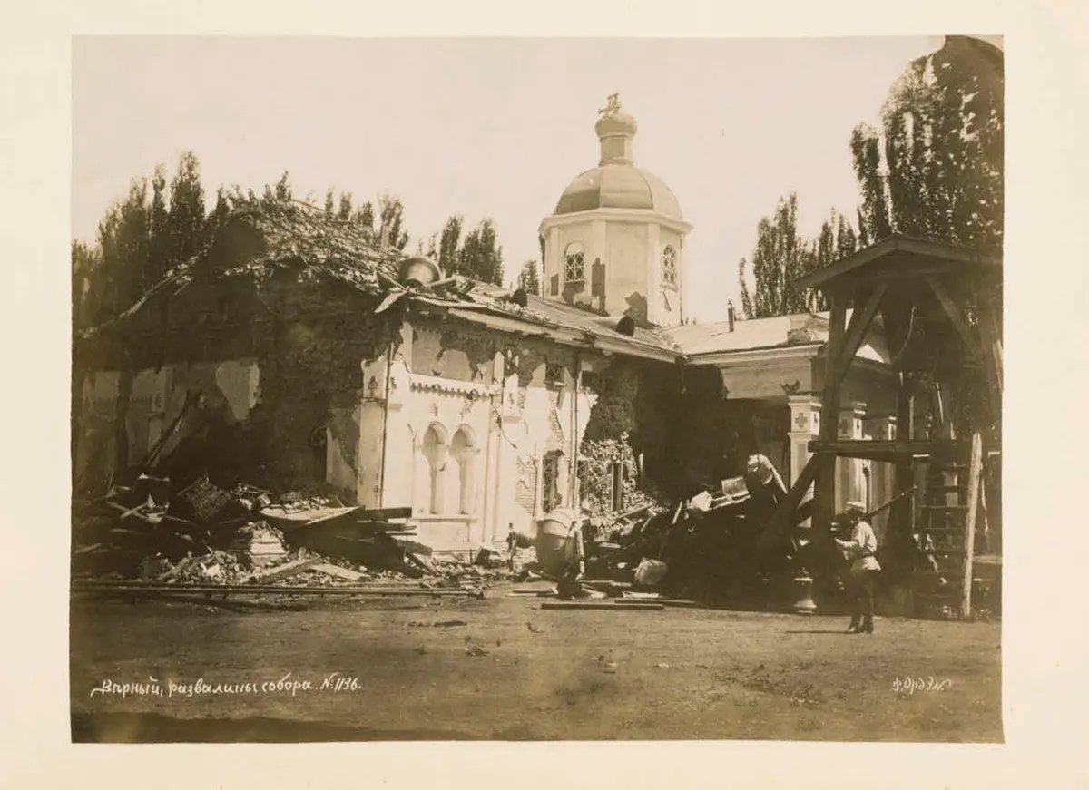 Fotografia de álbum da catedral arruinada de Verny, com a cúpula ainda de pé, escombros no pátio e uma pessoa junto à estrutura de madeira à direita.