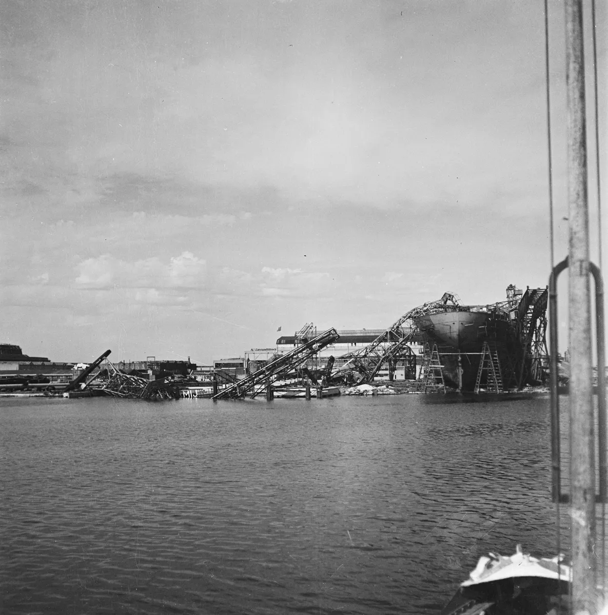 Foto em preto e branco com estruturas de guindastes destruídas junto à água e a proa de uma embarcação no porto.