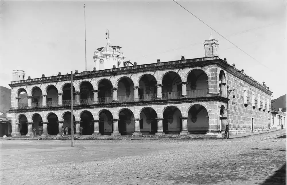 Fotografia em preto e branco do antigo Ayuntamiento de Antígua, prédio de dois andares com arcadas de pedra, colunas e torre com relógio, visto de uma rua de paralelepípedos.