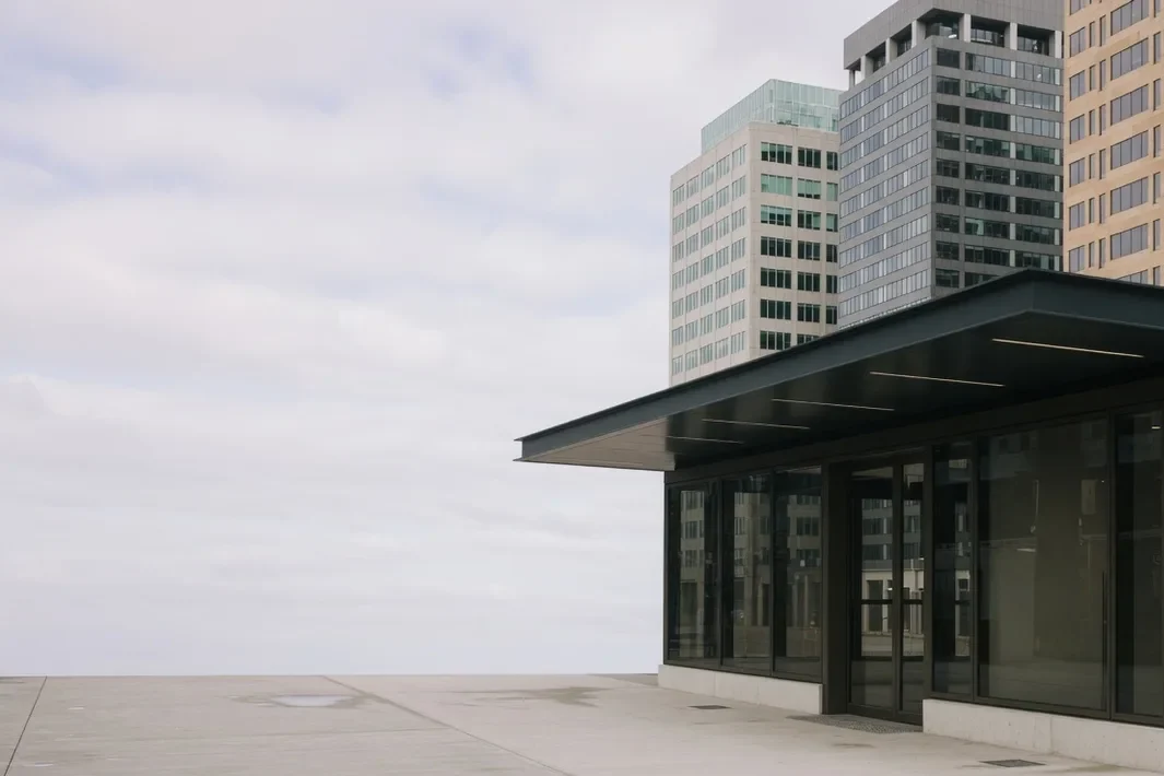 Entrada envidraçada de estação, com cobertura escura e torres ao fundo.