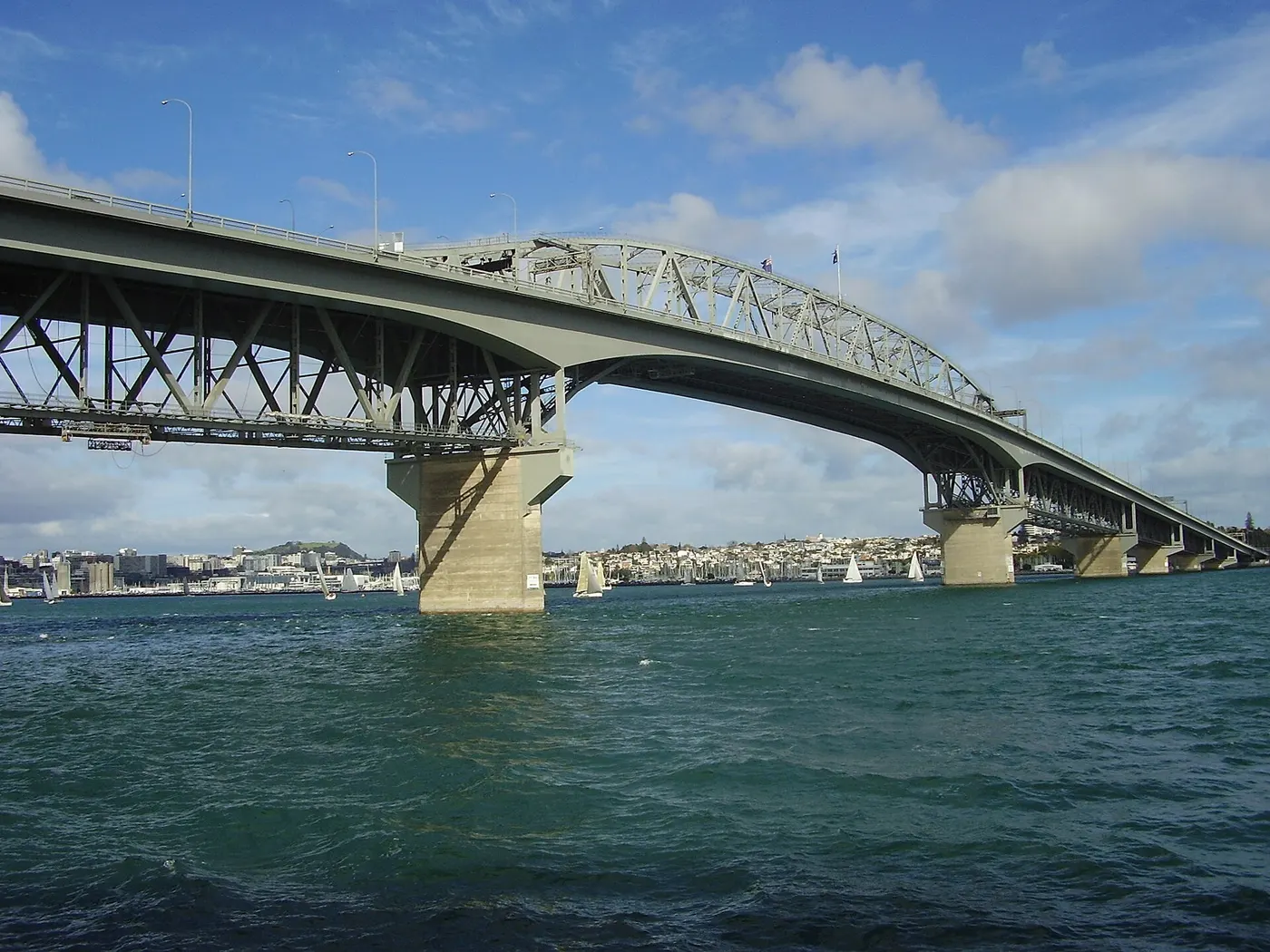Uma ponte de treliça de aço sobre a água, com pilares de concreto, veleiros e a cidade ao fundo.