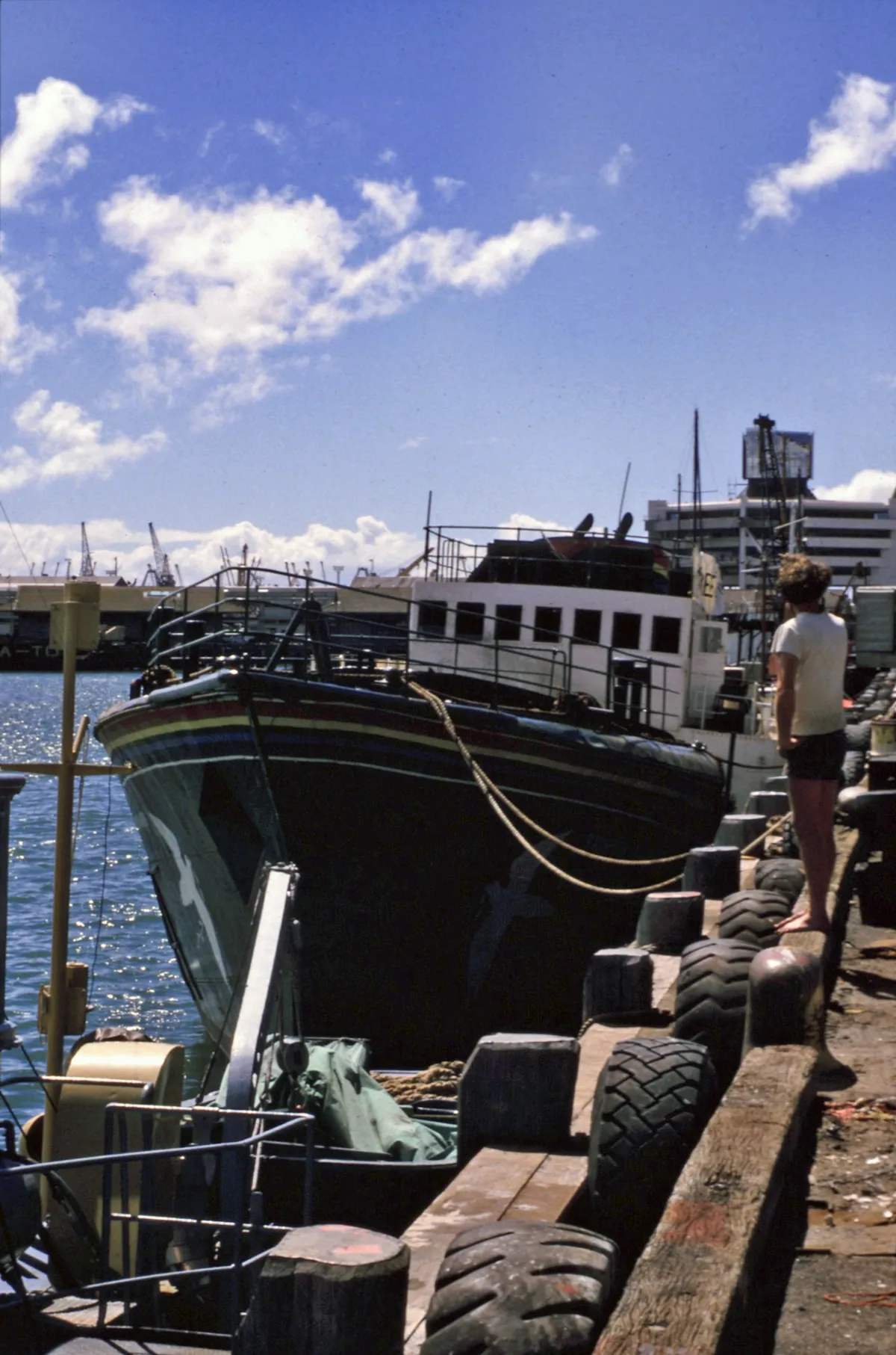 A proa escura de um navio no cais, com pneus ao longo do píer e uma pessoa vista de costas.
