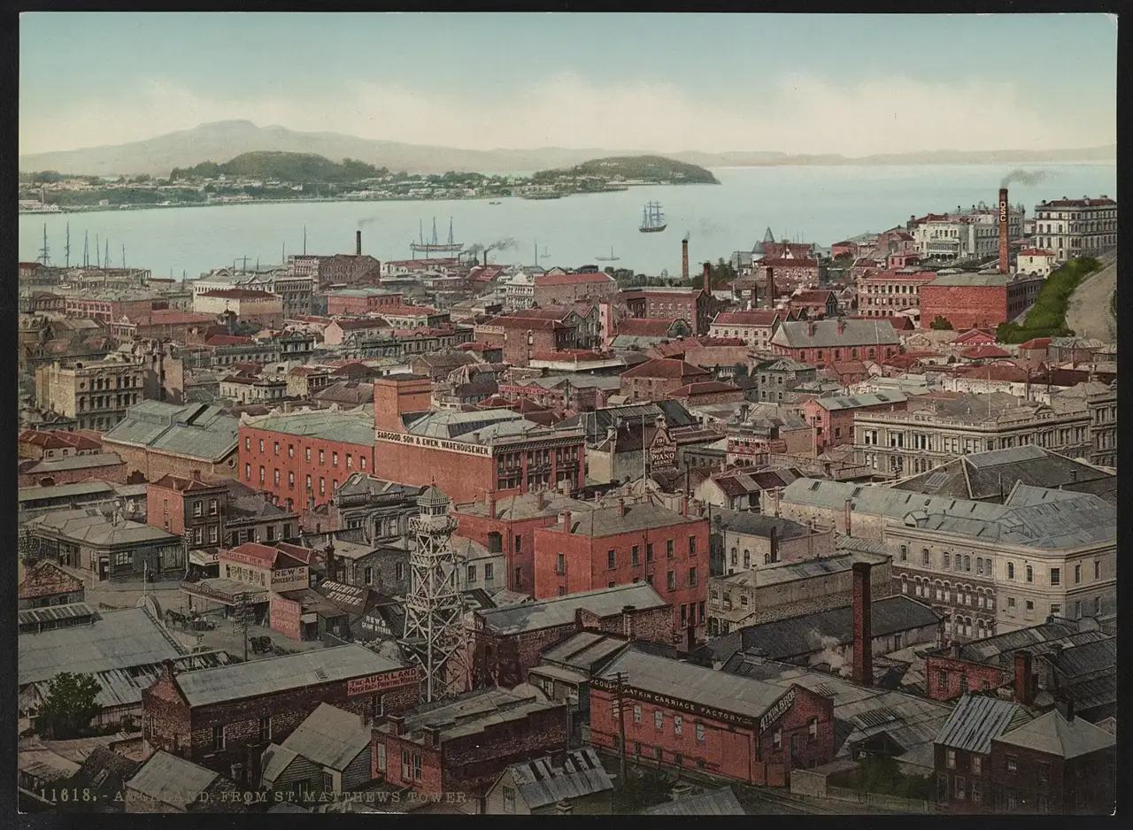 Fotocromo colorido dos telhados de Auckland vistos de cima, com o porto, navios à vela e a ilha vulcânica de Rangitoto no horizonte.