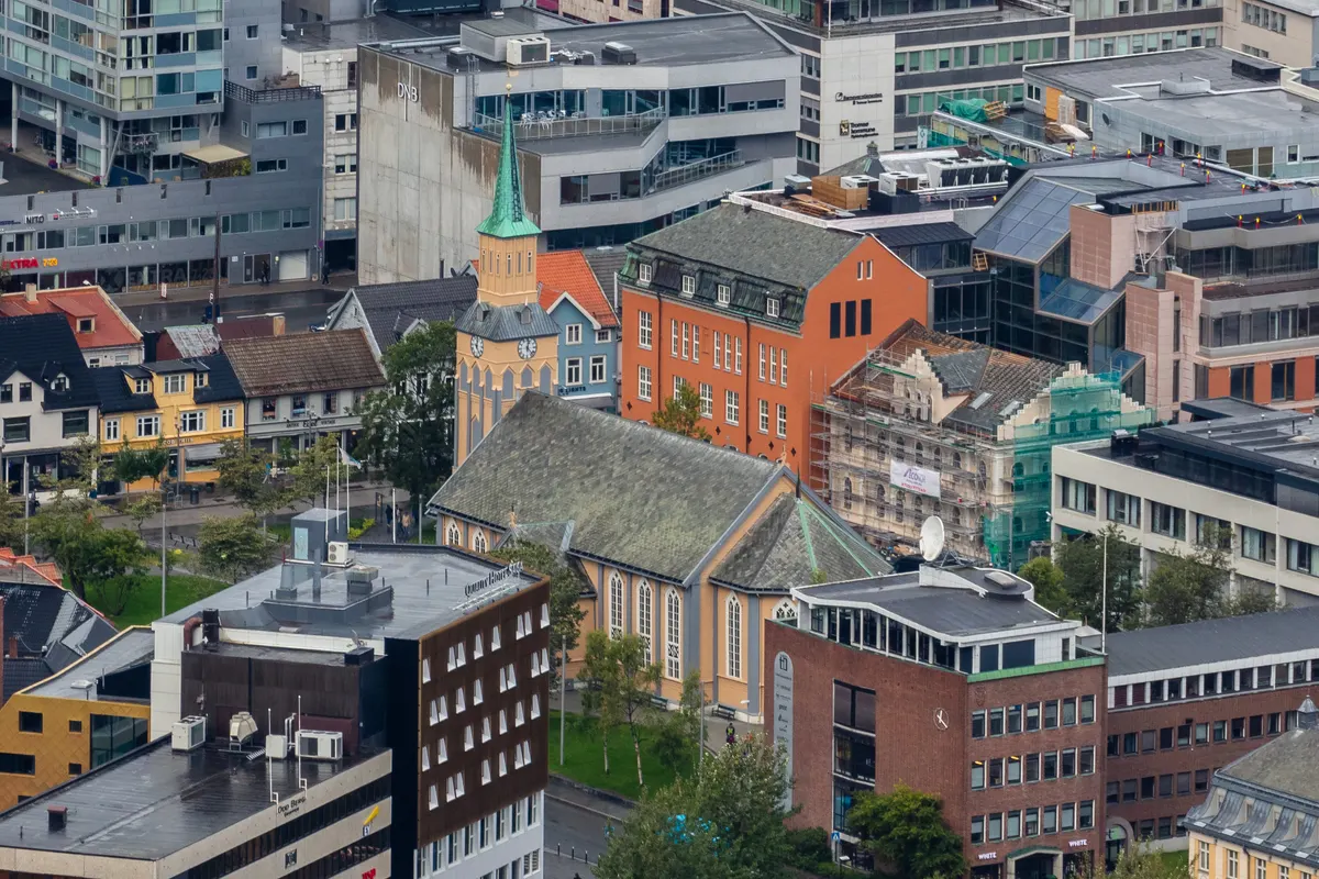 Catedral amarela com telhado cinza e agulha verde entre edifícios do centro, vista de um ponto elevado.