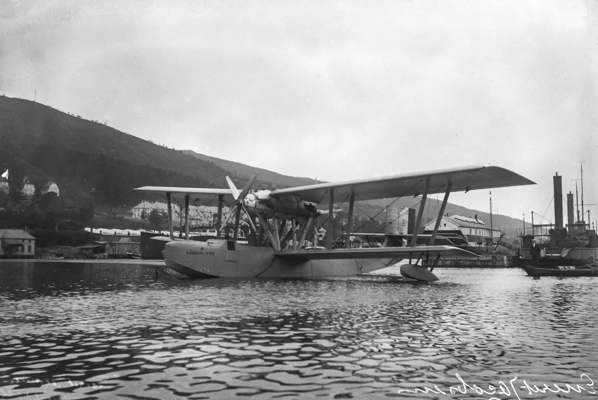 Hidroavião biplano sobre a água, com hélice, asas e flutuadores, edifícios e barcos ao fundo; marca manuscrita na borda inferior.
